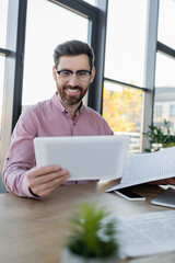 Happy businessman looking at digital tablet near papers and gadgets in office.