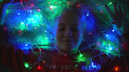 blonde teenager girl lies in garland lights for christmas, shows heart with a roar and smiles