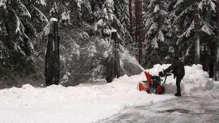 a man cleans snow with a snowblower on the road near a snowy winter forest