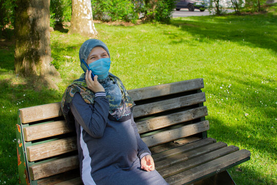 Young muslim pregnant woman wearing face mask talking on the phone while sitting in the park - Powered by Adobe
