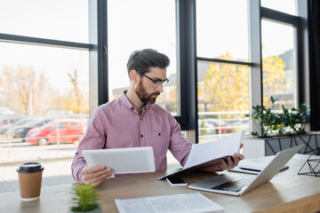 Businessman looking at papers and holding digital tablet near devices and coffee to go in office.