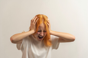 teen girl with red hair in a white t-shirt on a light background hold her head with her hands and scream