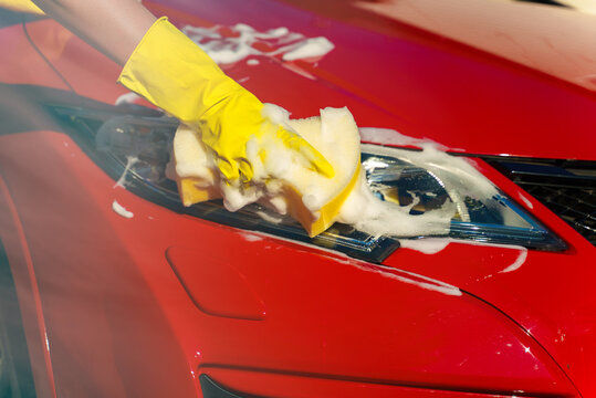 Woman in gloves washes car headlight with sponge.