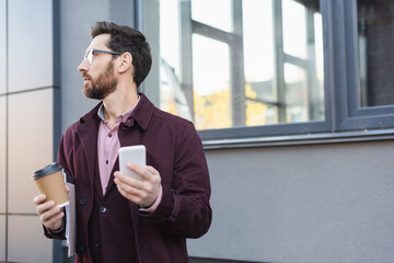 Side view of businessman with paper folder and coffee to go using smartphone outdoors.