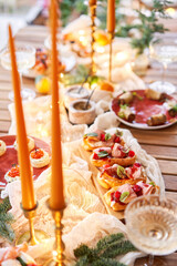 Christmas dinner feast. A small table is served with snacks, bruschettas, and canapes. A decorated dining table with champagne glasses, candles and christmas tree an garland in background