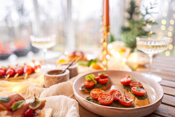 Christmas dinner feast. A small table is served with snacks, bruschettas, and canapes. A decorated dining table with champagne glasses, candles and christmas tree an garland in background