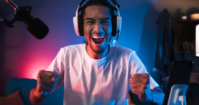 Young Asian man playing online computer video game, colorful lighting broadcast streaming live at home. Ecstatic celebration winning a match. Gamer lifestyle, E-Sport online gaming technology concept