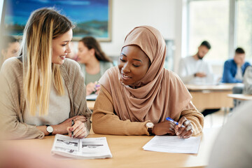 Happy Muslim student learning with her female Caucasian friend during class at the university. - obrazy, fototapety, plakaty