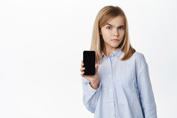 Fototapeta premium Little girl, teen child showing smartphone screen, mobile phone app display, smiling and looking happy at camera, standing over white background