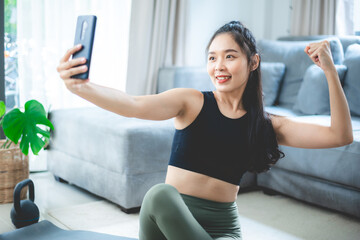 Fototapeta na wymiar Portrait of cheerful young Asian woman in sportswear showing biceps and muscles sitting on yoga mat and taking a selfie from smartphone at home after practicing yoga and exercise