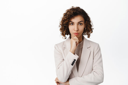 Confident young corporate woman, listening with thoughtful face, looking determined at camera, wearing suit, standing over white background