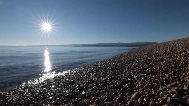 Water waves over small round pebbles on zlatni rat beach in croatia on a summer day with strong sun and lens flare in the background. Slow motion video