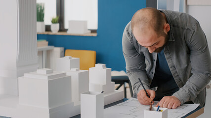 Close up of architect taking notes on blueprints plan to work on building model and maquette. Man builder using construction layout plans to design urban project for real estate development.