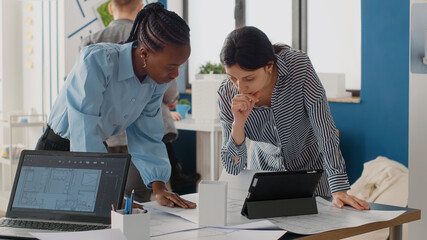 Close up of diverse women analyzing blueprints plan with digital tablet, doing teamwork for project. Architects working on construction layout and structure for urban development.