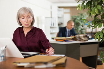 Portrait of busy focused aged female entrepreneur sitting at office desk with papers and laptop ..