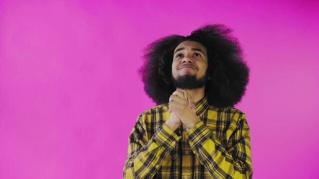 A young man with an African hairstyle on a pink background crosses his fingers and prays. Emotions on a colored background.