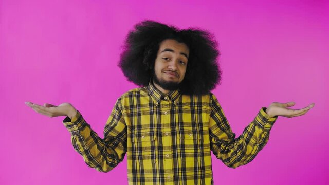 A young man with an African hairstyle on a pink background shrugs his shoulders. Emotions on a colored background.
