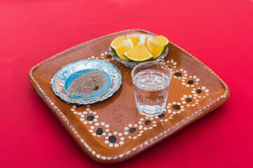Mezcal with worm salt and orange slices to accompany it. on a clay plate and a red background.