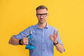 smiling scientist man in glasses hold microscope show ok gesture on yellow background, science