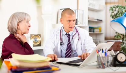 Senior woman visiting qualified doctor in medical office with complaining of sore throat. Focused therapist examining female patient and prescribing treatments while sitting at table - obrazy, fototapety, plakaty