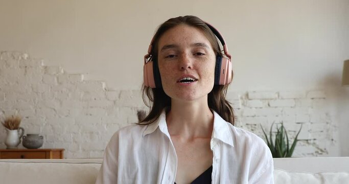 Young teenage girl wear headphones sit on sofa start distancing conversation via video conference, webcam head shot portrait. Seeker passing job interview use modern tech, video call and app concept
