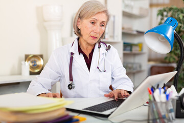 Focused professional aged female physician filling up medical forms on laptop while sitting at...