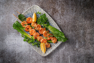 Grilled king prawns with lemon and herbs on a plate against a gray stone table, fish kebab