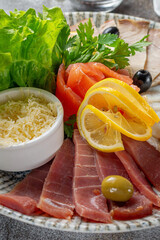 An appetizer in a restaurant, assorted meats. Sliced meat and sausage in layers on a plate with greens against a gray stone table 