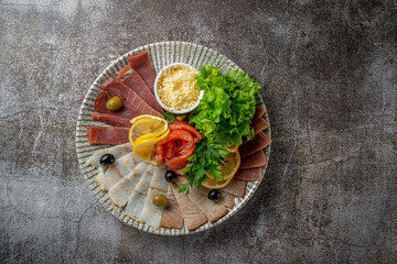 An appetizer in a restaurant, assorted meats. Sliced meat and sausage in layers on a plate with greens against a gray stone table 