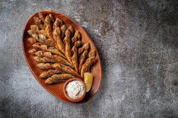An fish appetizer in a restaurant, fried sprat on a wooden plate with lemon and cream sauce against a gray stone table 