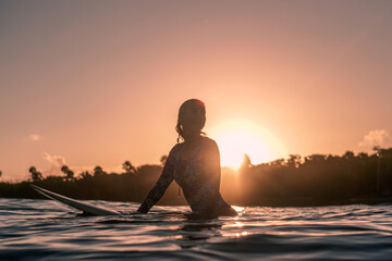 Portrait of blond surfer girl on white surf board in blue ocean pictured from the water at golden sunrise time in Encuentro beach - obrazy, fototapety, plakaty
