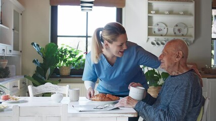 Young female caregiver or healthcare worker visiting senior man indoors at home, assisting with breakfast