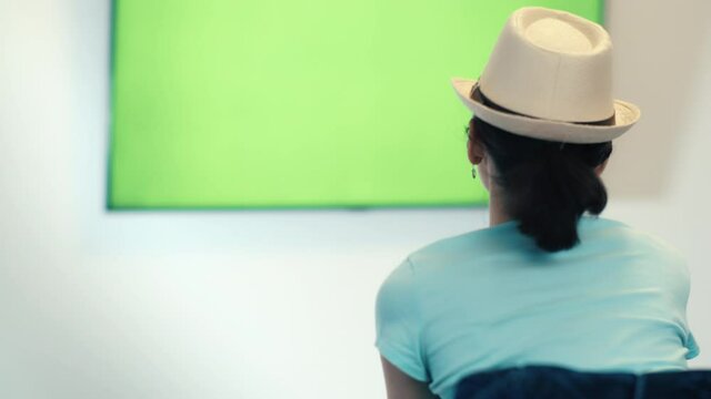 Brunette Girl in a hat Sits in Font of a Geen Sreen TV, Switches Channels, Green Chroma Key Screen TV, Relaxing on a Sofa, Attentively Watches Information on TV