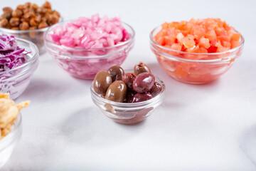 A view of a glass condiment bowl of Kalamata olives, among a variety of other chopped ingredients.