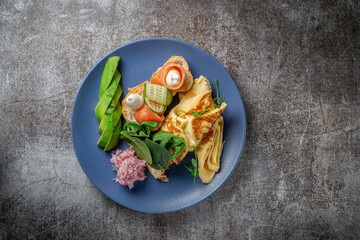 Omelet with salmon, fresh herbs and tomatoes in a blue plate with spices against a gray stone table .