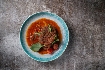 Beef stew with broth and herbs in a blue plate with spices and black pepper against a gray stone table .