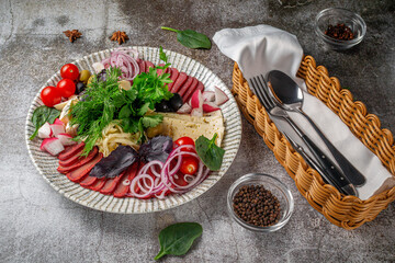 An appetizer in a restaurant, assorted meats. Sliced meat and sausage in layers on a plate with greens against a gray stone table