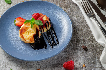 Fried pancakes with chocolate syrup and raspberries and mint in a blue plate against a gray stone table. A sweet breakfast in the morning