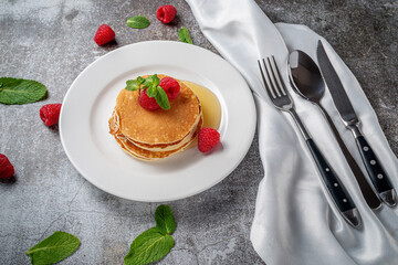 A delicious breakfast. Sweet fried raspberry and mint pancakes in a white plate with sour cream on a gray stone table with a napkin and cutlery