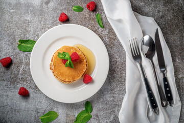 A delicious breakfast. Sweet fried raspberry and mint pancakes in a white plate with sour cream on a gray stone table with a napkin and cutlery