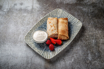 Obraz na płótnie Canvas A delicious breakfast. Sweet fried pancakes filled with strawberries in a white plate with sour cream on a gray stone table with a napkin and cutlery