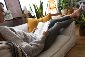 Happy young caucasian woman reading a book at home. Relaxing on the sofa.