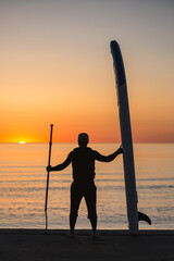 A surfer stands on the ocean shore holding a SUP board and a paddle in his hand. Standup...