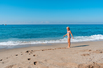 Blond preschool girl wearing colorful swimsuit near the sea on the sand beach on a beautiful summer sunny day.