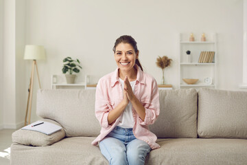 Online webcam view of happy adult woman sitting on sofa with notebook at home, smiling at camera and rubbing her hands in anticipation while waiting for her friend to show her surprise on video call