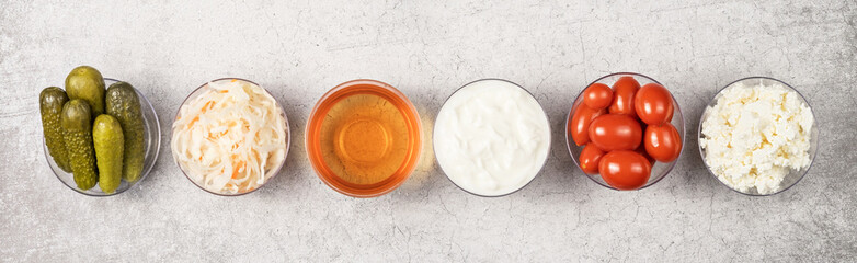 A variety of fermented foods for gut health. Bowls on a gray background. Cucumbers, tomatoes, sauerkraut, yogurt, cottage cheese, apple cider vinegar. Banner.
