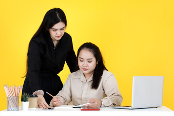 Studio shot of Asian happy female mentor employee standing smiling  help teaching job training to chubby trainee colleague sitting working with paperwork document at office desk on yellow background