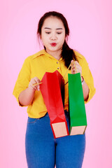 Portrait studio shot of Asian young happy chubby plump female shopaholic shopper wearing casual outfit standing carrying opening colorful shopping bags smiling  on pink background