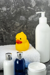 Obraz na płótnie Canvas baby bathing products, baby care, yellow rubber duck for playing in the bath. Children's cosmetics, toys and a white towel on the table on a gray background. Selective focus.