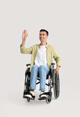 Young man in wheelchair waving hand on light background